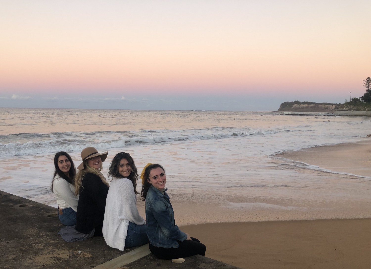 Group of four students sitting on a ledge by the ocean at sunset