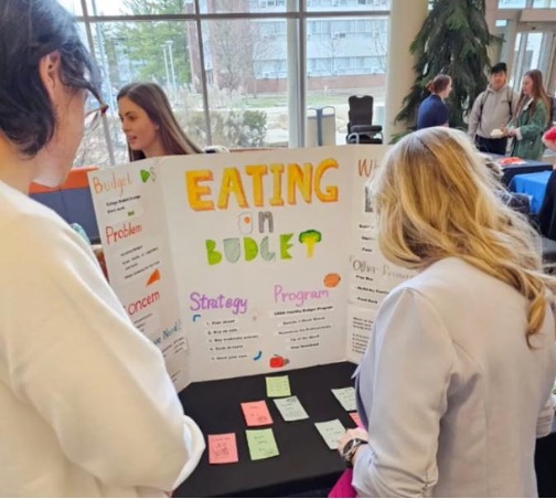 two people standing in front of a table with a standing cardboard poster that reads eating on budget