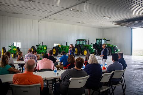 Picture of Janet Kreig speaking to attendees seated at tables in a machine shed with John Deere tractors in the background.