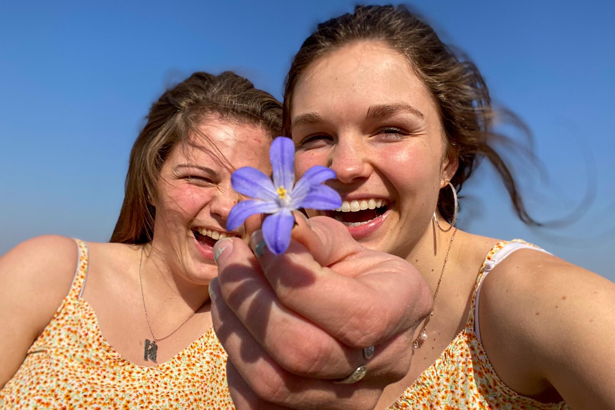 Sisters, Jamie and Jennie, smile together while holding out a small, purple flower. 