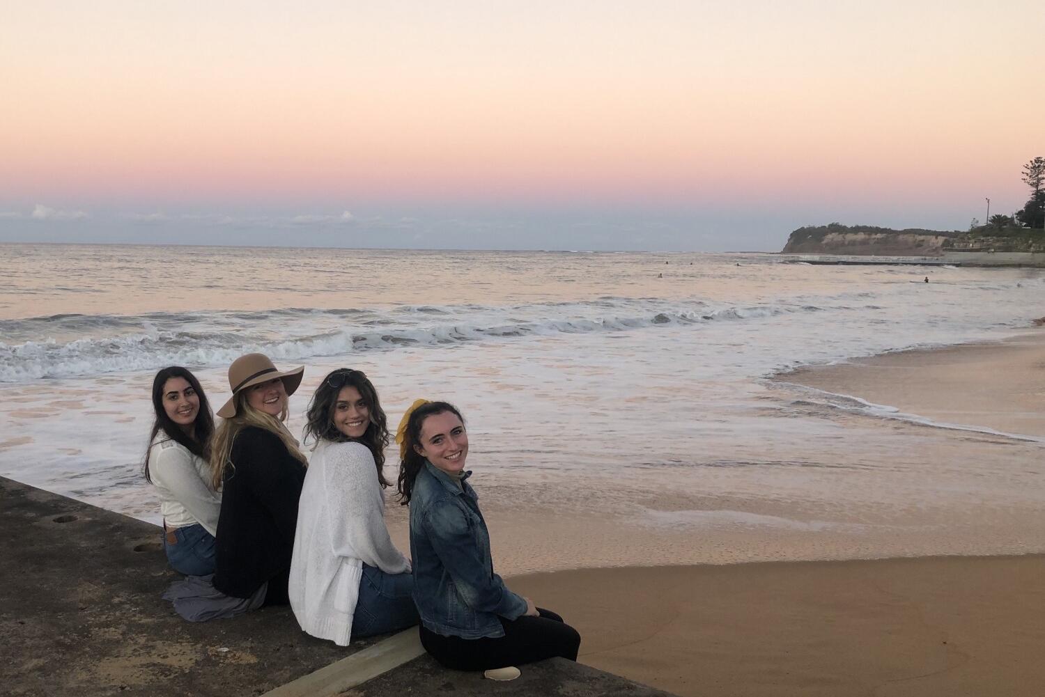 Group of four students sitting on a ledge by the ocean at sunset