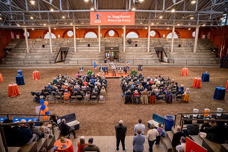 A view of the ceremony looking towards the stage flanked by balloons and prints of the arena’s bronze plaques, showing ground-level seats and stadium seating filled with attendees; the large orange “Dr. Doug Parrett Memorial Arena” banner overhead was unfurled during the ceremony. 