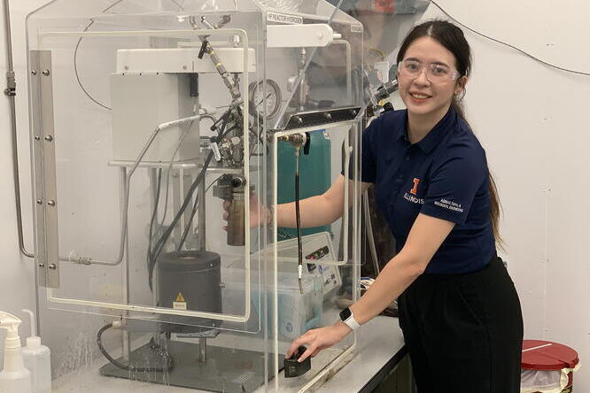 Sabrina Summers, University of Illinois, demonstrates hydrotreating biocrude oil from food waste. A woman in a blue t-shirt and safety glasses demonstrate lab equipment