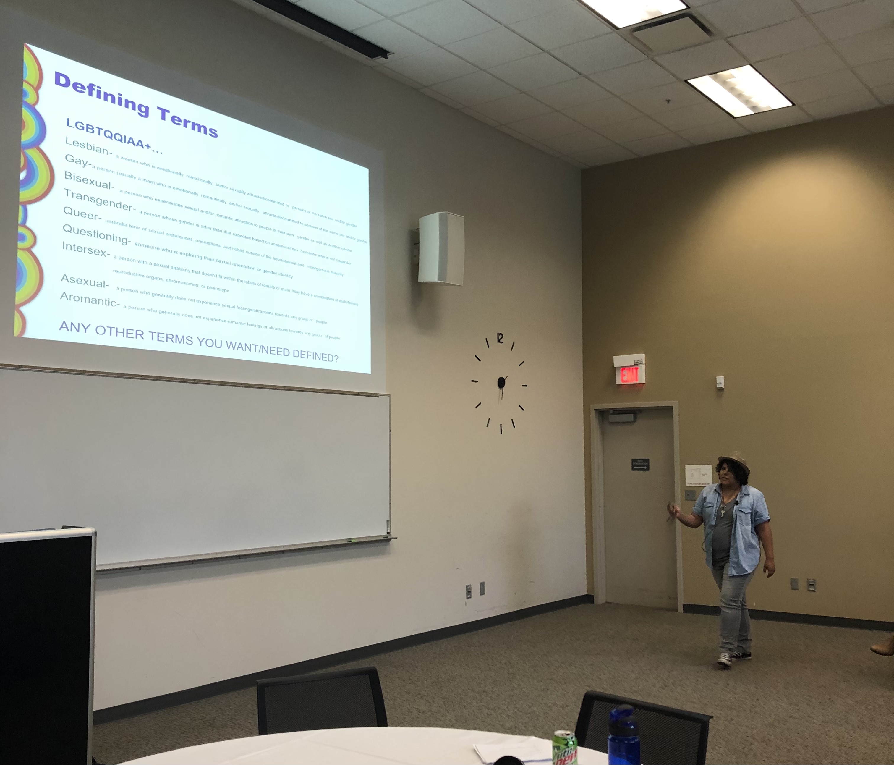 A person stands near a wall, presenting a slide titled "Defining Terms" that lists and defines various LGBTQIA+ identities in a classroom setting.