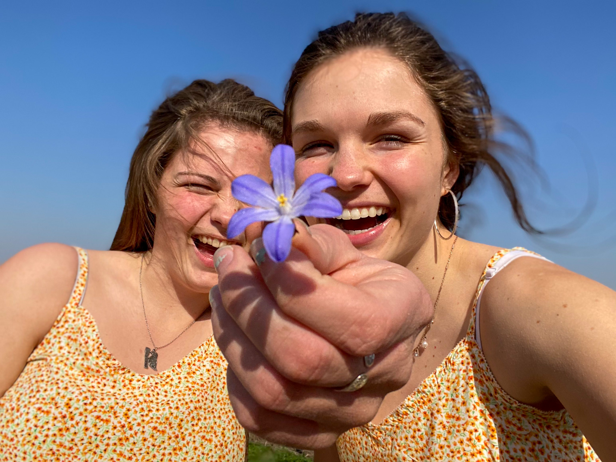 Sisters, Jamie and Jennie, smile together while holding out a small, purple flower. 