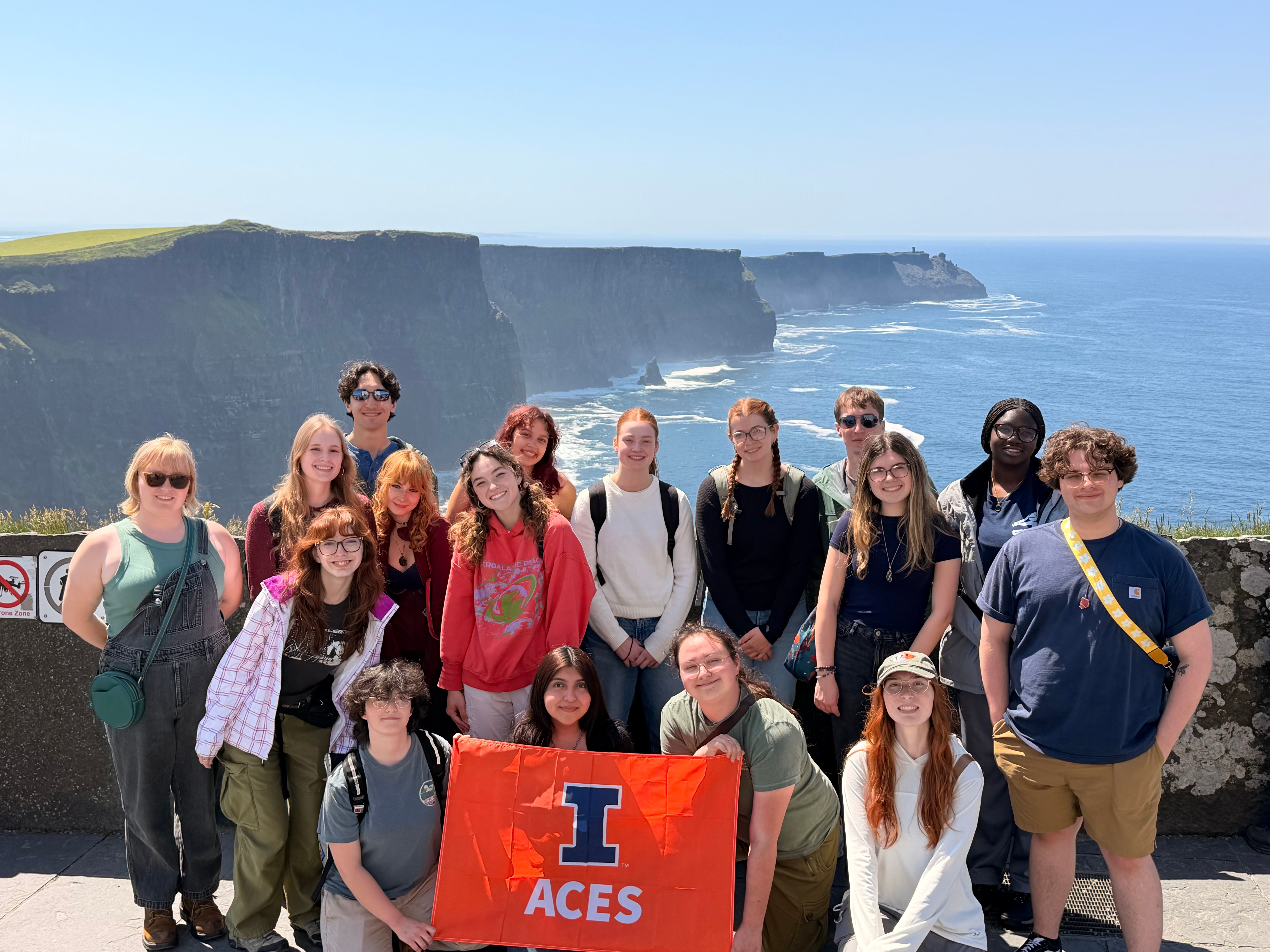 A group of students in the foreground hold an orange banner with the ACES logo. Behind them, the ocean appears below large cliffs.