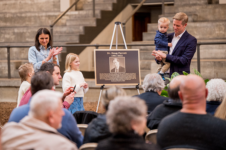 Doug Parrett’s son John Parrett (right) along with his wife Crystal (left) and children stand next to a print of the bronze plaque titled Dr. Doug Parrett 1951-2022.