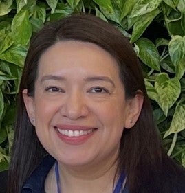 Diana Rodriguez is a Latina woman, with straight, dark brown hair that falls just past her shoulders. She is wearing a blue shirt and smiling broadly in front of green leaves. 