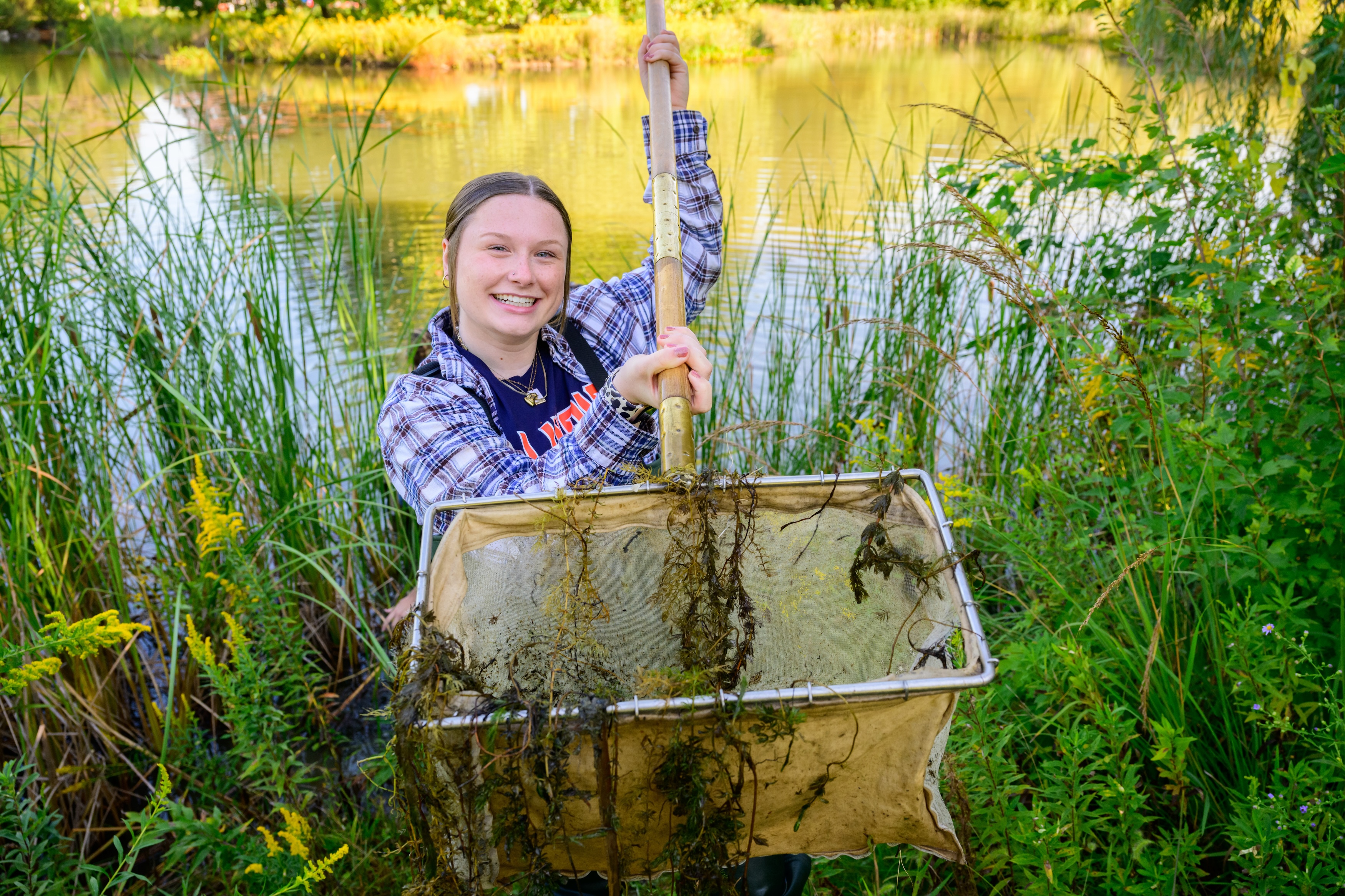 A smiling person wearing a plaid shirt holds up a large net filled with aquatic plants by a lakeside surrounded by tall grasses and greenery.