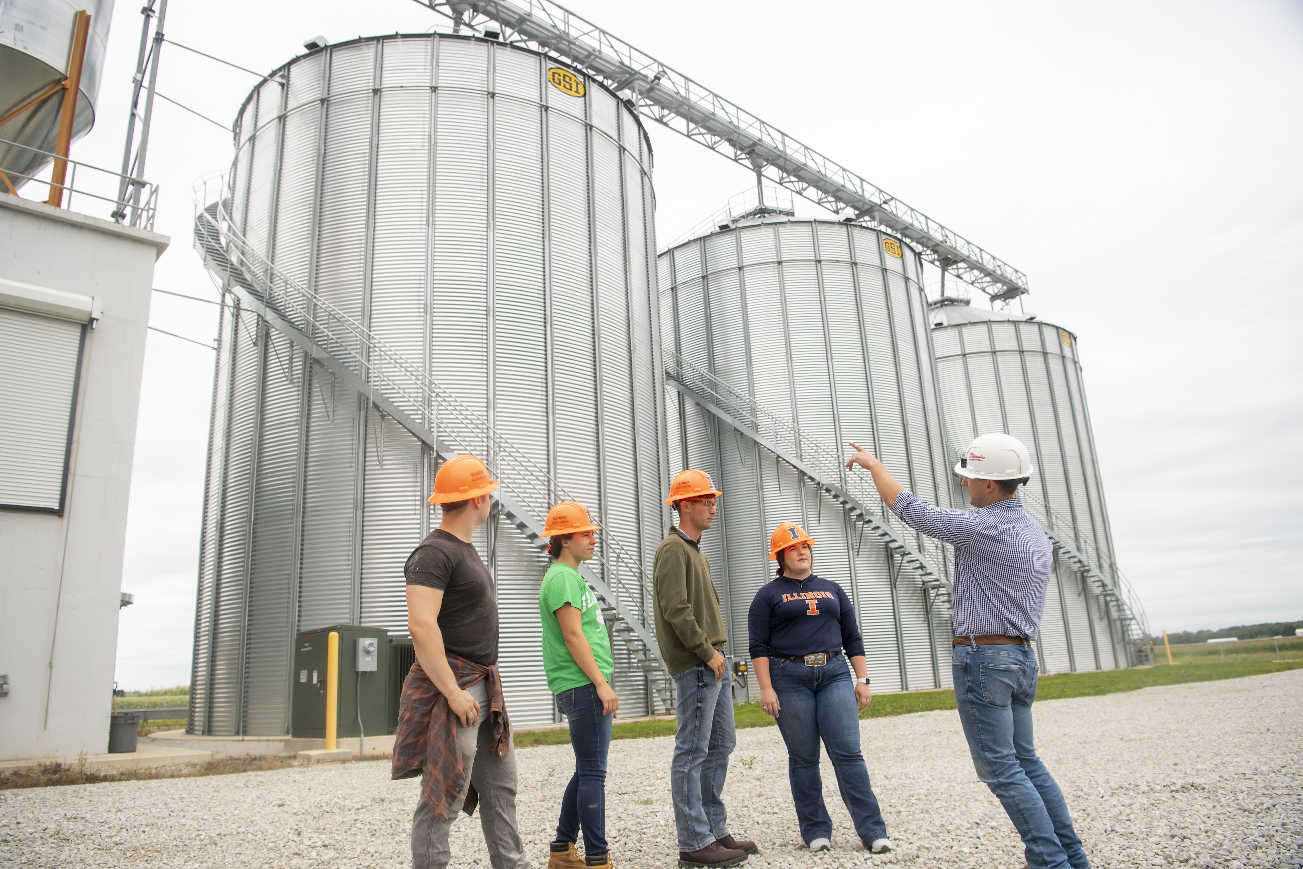 A group of people wearing hard hats stand near large grain silos, with one person gesturing and explaining something to the others.