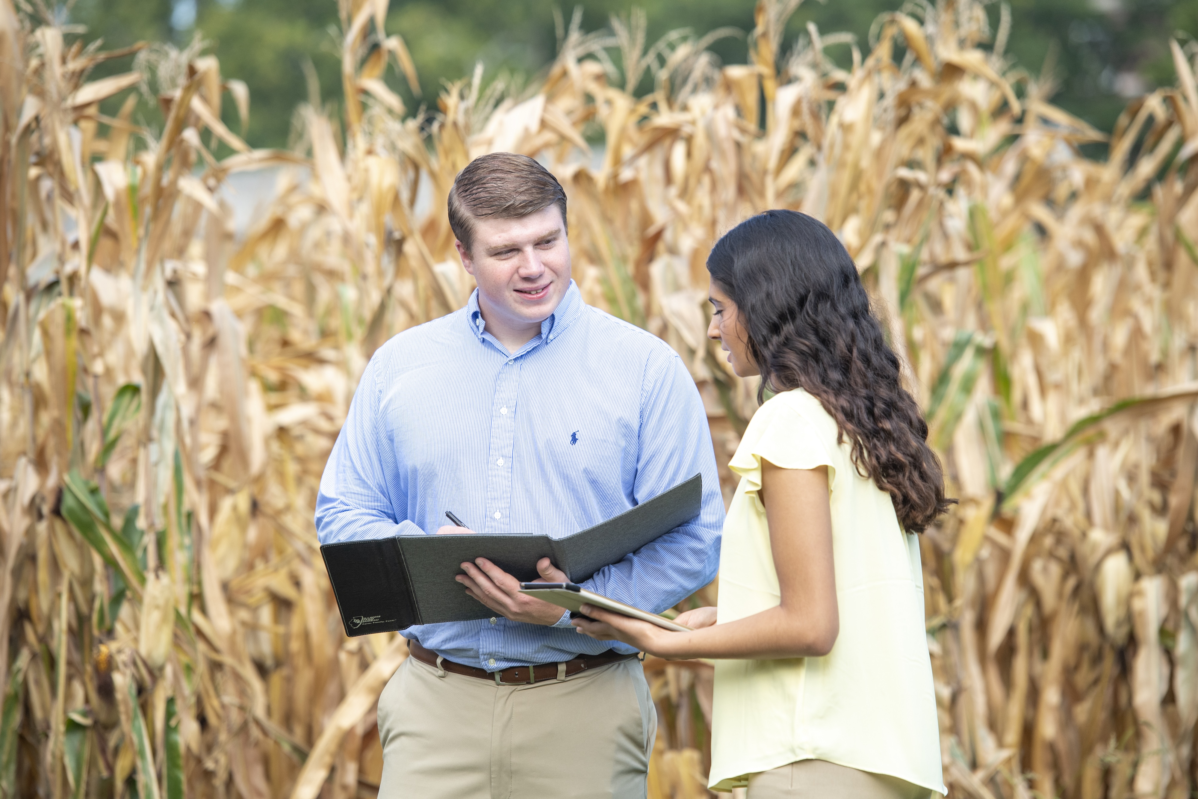 Two people stand in front of a cornfield, looking at documents in a folder and tablet, engaged in discussion.