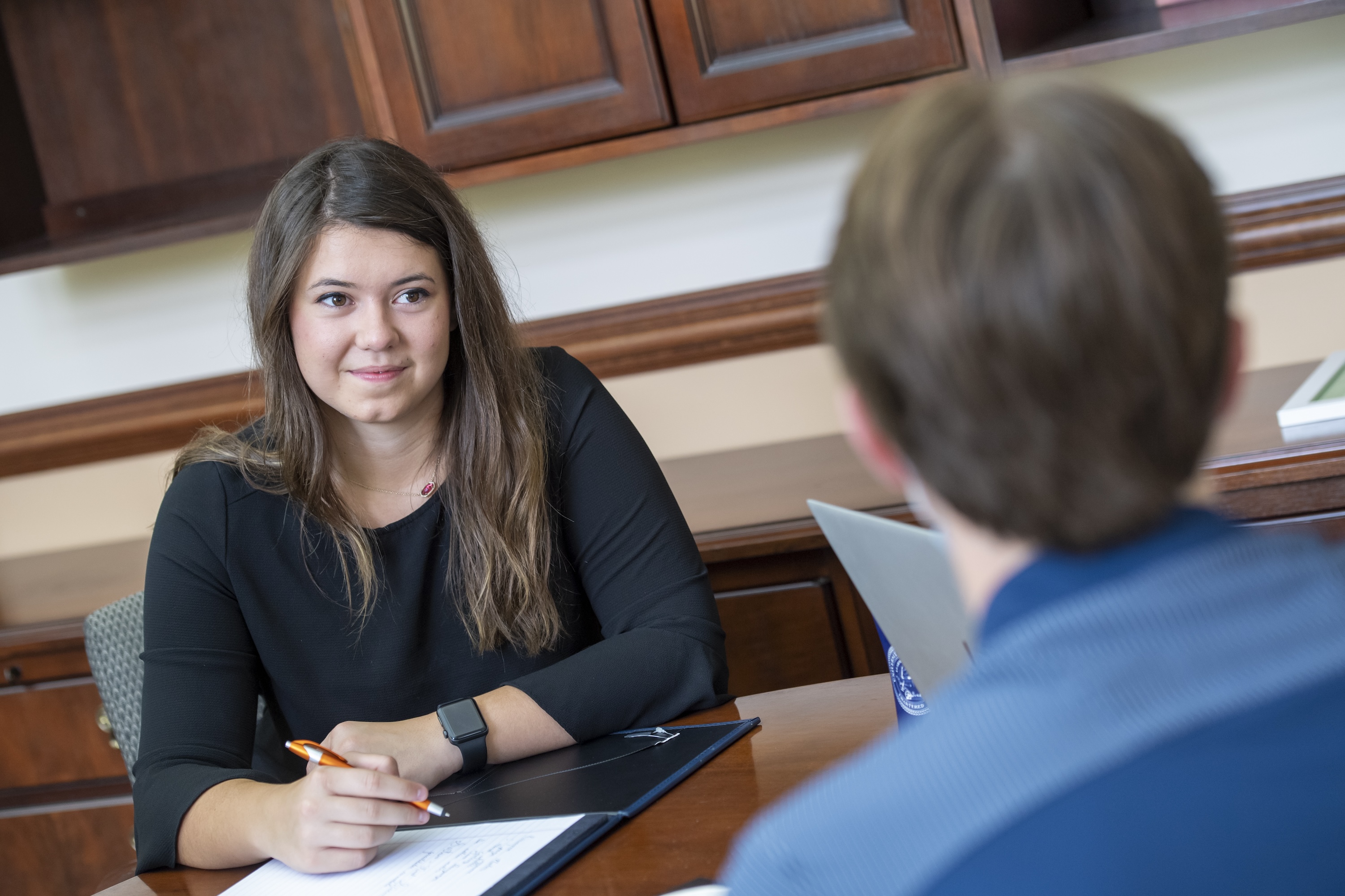 A young woman attentively listens during a meeting, holding a pen and notepad, while seated across from another person.