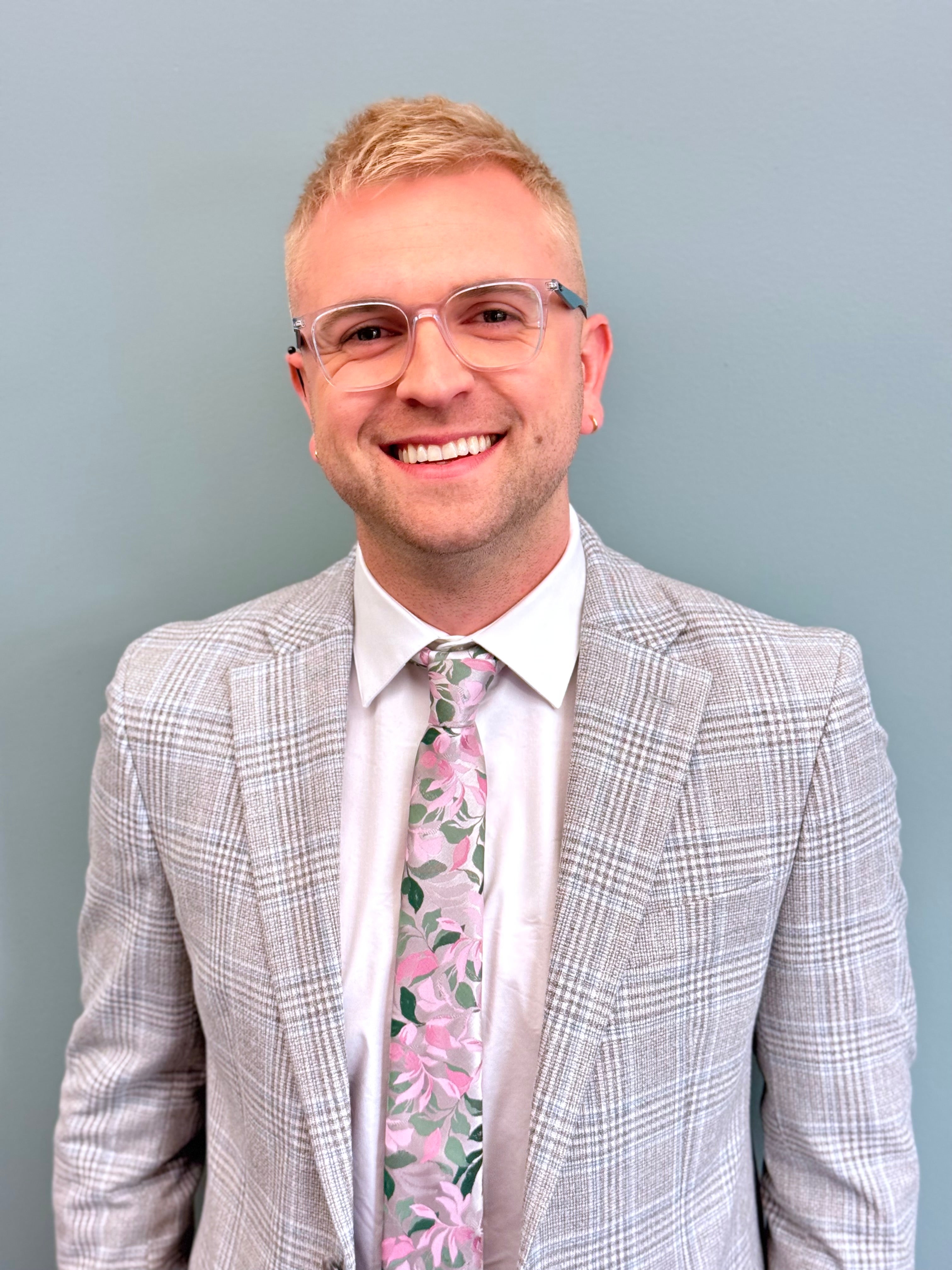 Matthew Rivas-Koehl is a white man, with blonde hair, wearing glasses and a gray jacket, white button up shirt, and floral tie. 