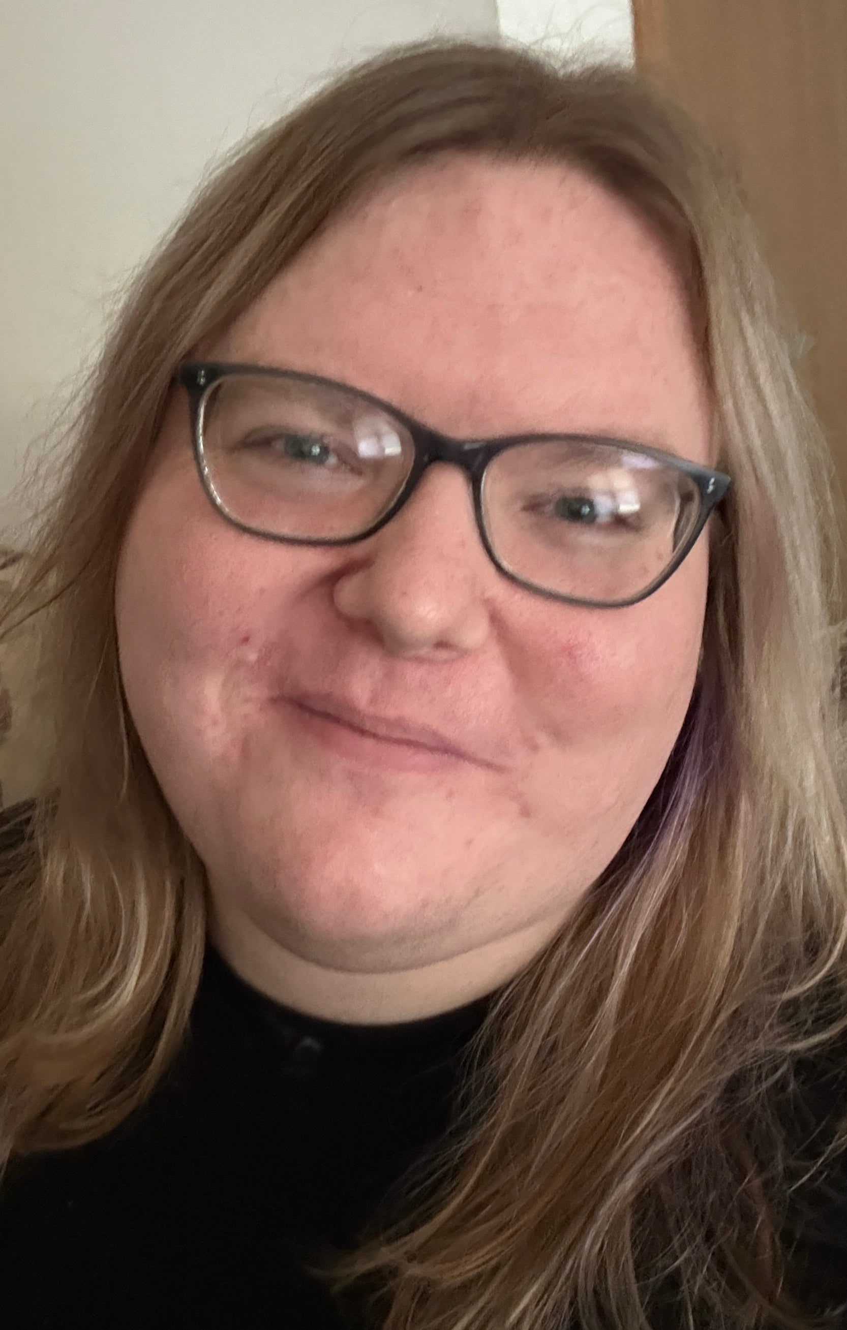 Mattie is a white individual, with dark blonde hair past her shoulders. She is wearing black-framed glasses and is smiling at the camera. 