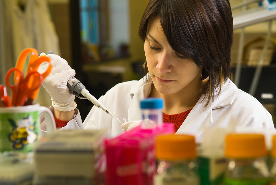 A scientist in a lab coat uses a pipette while working at a lab bench surrounded by colorful laboratory equipment.