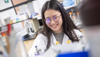 A person wearing glasses and a lab coat smiles while working in a laboratory.