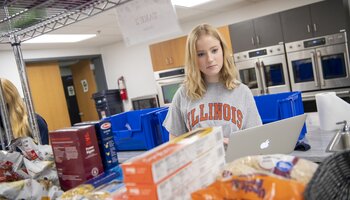 A person wearing an Illinois shirt uses a laptop surrounded by various food items and boxes in a kitchen setting with ovens in the background.