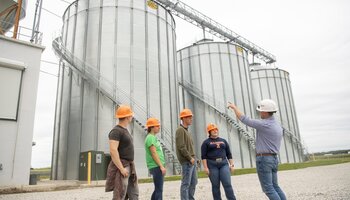 A group of people wearing hard hats stand near large grain silos, with one person gesturing and explaining something to the others.