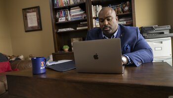  A man in a suit sits at a wooden desk working on a MacBook laptop, with a bookshelf and printer in the background. A blue mug and a few papers are on the desk.