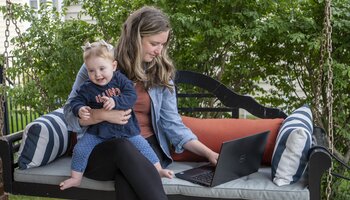 A woman holding her child while sitting and working on a laptop.