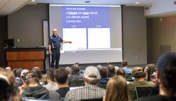 A man stands in front of a classroom, presenting a slide about trading concepts to a group of students seated with laptops.