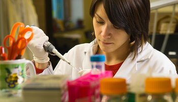 A scientist in a lab coat uses a pipette while working at a lab bench surrounded by colorful laboratory equipment.