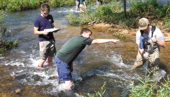 Three people stand in a shallow creek, one holding a measuring device, another pointing, and the third taking notes on a clipboard.