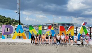 A group of people sit in front of large, colorful letters spelling "PANAMÁ" at a marina, with boats and the city skyline visible in the background under a cloudy sky.