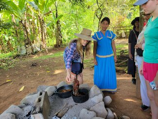 A group of people gathers outdoors as one person tends to food cooking over a fire, while a woman in a bright blue traditional dress observes nearby, surrounded by lush greenery.