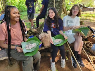 Three people sit outdoors, smiling and holding food served on large banana leaves while surrounded by trees and nature.