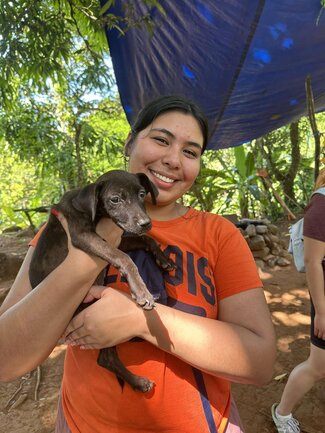 A smiling person wearing an orange Illinois shirt holds a small black animal while standing outdoors under a tarp with trees in the background.