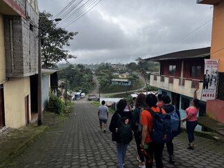 A group of people walk down a cobblestone street in a rural town, with houses on either side and a hilly, forested landscape in the distance under a cloudy sky.
