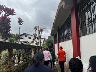 A group of people walk alongside a building with red columns, while decorative plants and houses are visible in the background under a cloudy sky.