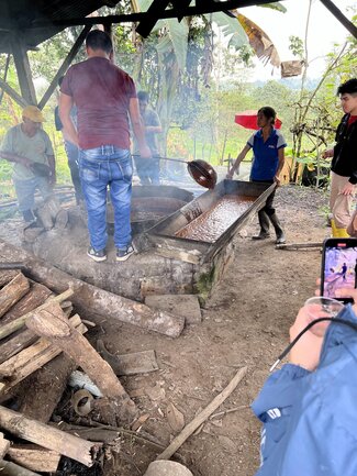 A group of people gather around an outdoor setup, where individuals are stirring a large vat of liquid over a fire, with wood logs and smoke in the foreground and tropical vegetation in the background.