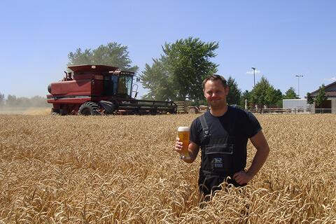 Matt Riggs stands in a wheat field holding a glass of Riggs Beer with a combine in the background.