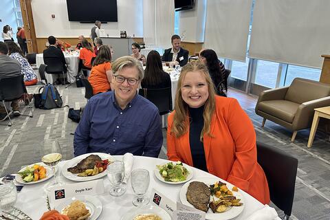 Two people in orange and blue sitting at a formal table. 