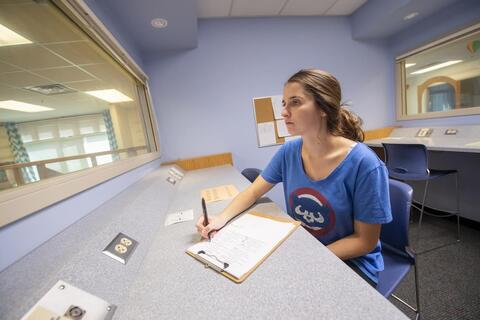 A young woman wearing a blue shirt sits in a small room with windows overlooking two classrooms