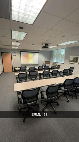 An empty classroom, labeled "ERML 138," is set up with rows of chairs and tables facing a projector screen displaying a presentation slide.