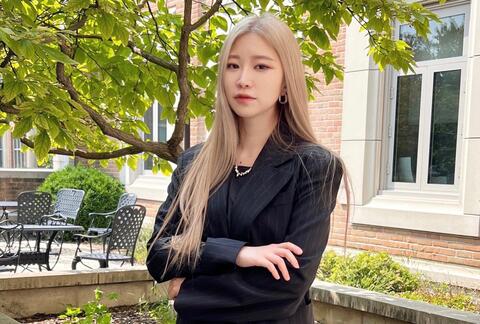 A young woman standing with her arms crossed in front of a building with a tree in the background