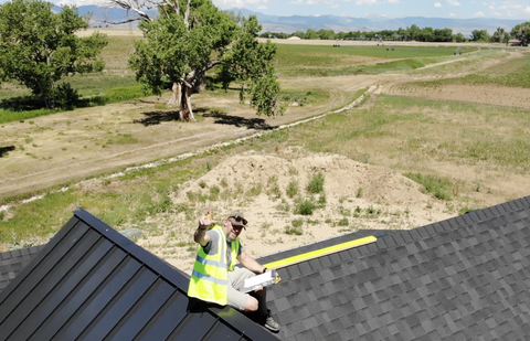 John Miller sits on the roof of a building installing solar panels and holding a yellow tool and waving, with open fields and a tree in the background
