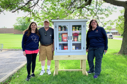 Three people flank a small outdoor pantry with trees and grass in the background