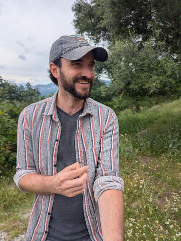 Man with a beard and wearing a hat stands outdoors. Trees and grasses are in the background.