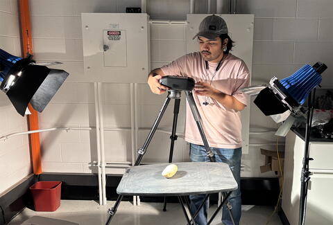 A man in a laboratory setting is holding a camera over a sweet potato on a table