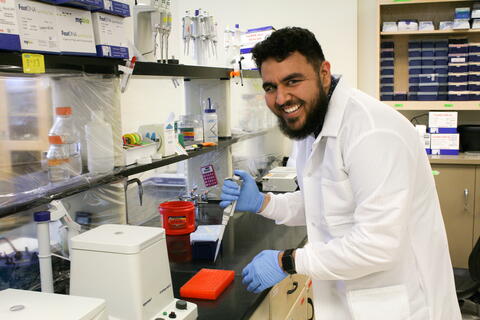 A bearded man wearing a white lab coat and blue gloves works with a pipette in a laboratory setting