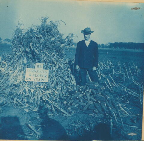 An old cyanotype photo of a man in a suit and hat standing next to a pile of corn with a sign