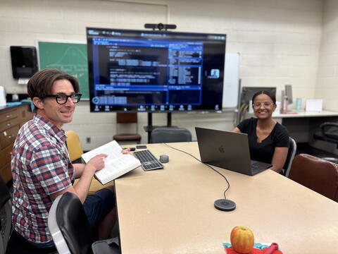 A man and woman sit at a table in a laboratory space. The man is holding an open book and the woman is in front of an open laptop. Behind them is a large screen filled with computer code.