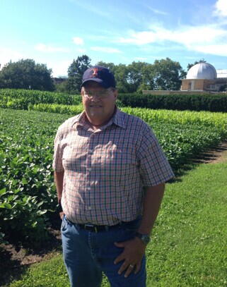 Man in baseball hat stands in front of a small field of soybeans. In the background, the University of Illinois observatory building appears.