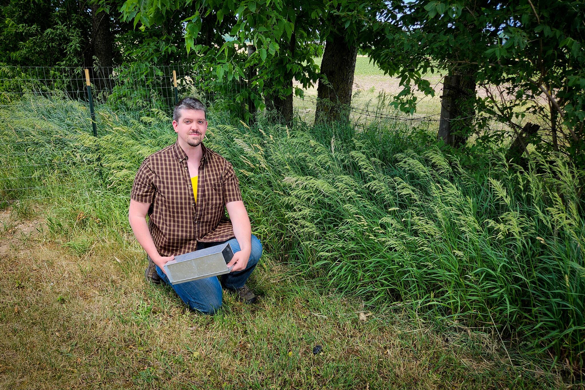 Nathan Alexander kneels outdoors holding a small mammal Sherman live trap.