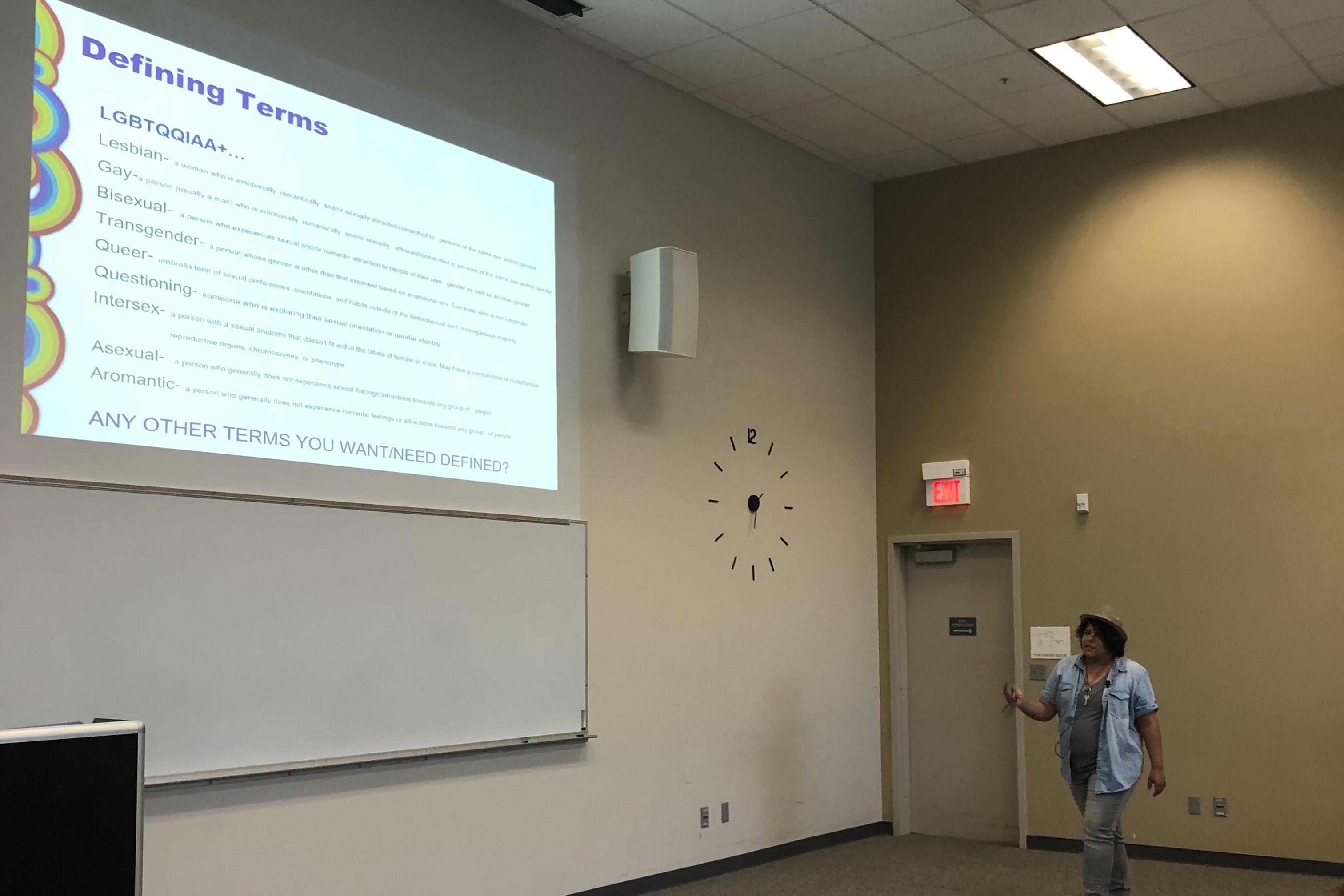 A person stands near a wall, presenting a slide titled "Defining Terms" that lists and defines various LGBTQIA+ identities in a classroom setting.