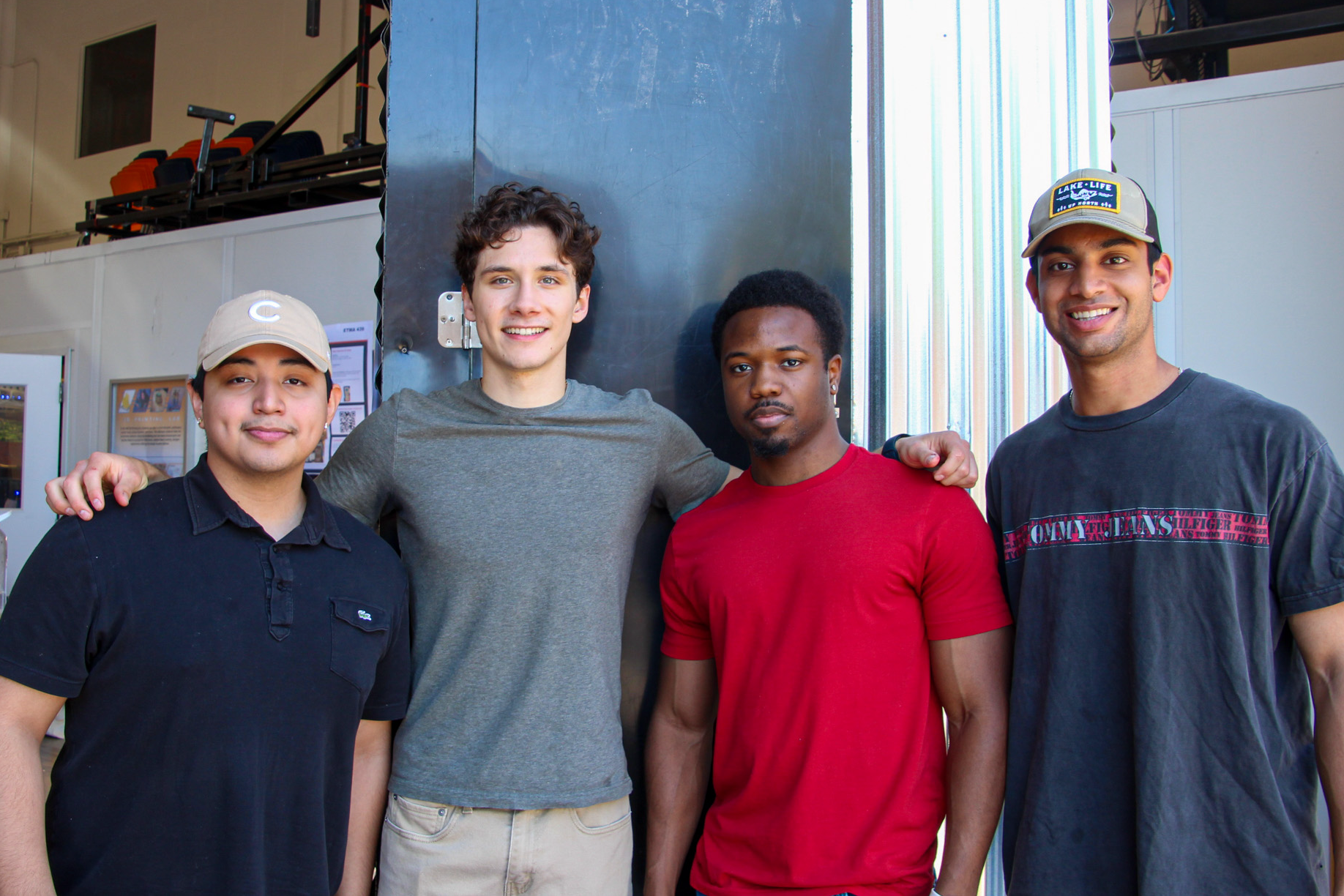 Four young men standing together and smiling in a workshop environment.